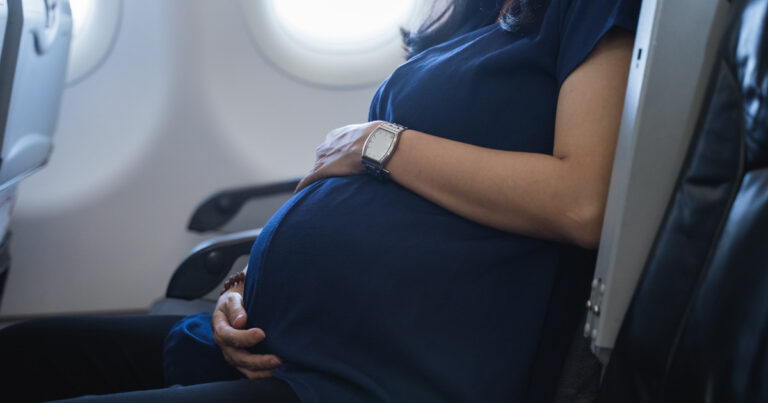 Avión en vuelo, ilustración de un nacimiento a bordo y símbolos de banderas representando la duda sobre la nacionalidad.